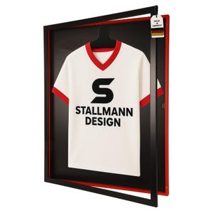 Op de afbeelding: Een wit t-shirt met rode randen en de tekst "STALLMANN DESIGN" in zwart, gepresenteerd in een zwart frame met een rode rand. Het shirt hangt aan een zwarte hanger. Een label met "MADE IN GERMANY" is aan het frame bevestigd.