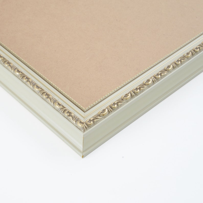 K&ouml;nnte beinhalten: Ein wei&szlig;er Bilderrahmen mit einem goldenen, verzierten Design am Rand. Der Rahmen ist aus Holz und hat eine matte Oberfl&auml;che.