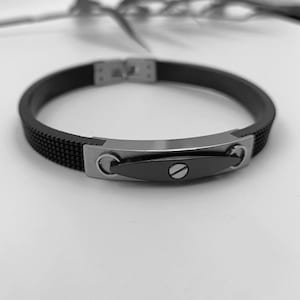 Puede incluir: Una pulsera de silicona negra con una placa de metal negro y un tornillo plateado. La placa tiene una forma redondeada y lisa.