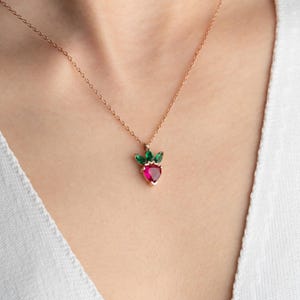 May include: A gold-colored necklace featuring a pendant shaped like a strawberry. The strawberry pendant has a red gemstone center with green gemstone leaves. The necklace is worn on a person wearing a white top.