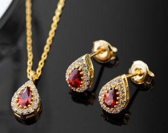 May include: A gold-coloured jewellery set comprising a necklace and matching earrings. The necklace features a teardrop-shaped pendant with a red gemstone centre, encircled by small, clear stones. The earrings echo the pendant design. The set is presented on a reflective black surface.