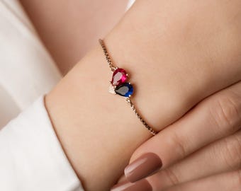 May include: A delicate gold bracelet with a heart-shaped charm featuring two teardrop-shaped gemstones, one red and one blue.