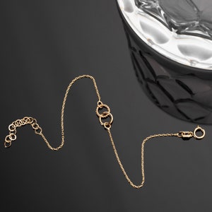 May include: A delicate gold necklace with a fine chain and a series of small, interconnected rings. The necklace features a clasp and an adjustable chain section. The jewelry is displayed on a reflective black surface.