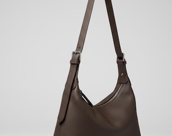 May include: A brown leather shoulder bag with a long strap and a zipper closure. The bag has a textured surface and a minimalist design. The strap is adjustable with a silver buckle.