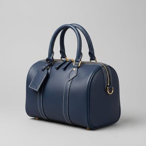 Op de afbeelding: Een marineblauwe leren duffeltas met goudkleurige hardware en witte stiksels. De tas heeft twee handvatten aan de bovenkant, een ritssluiting en een klein label. Een klassieke stijl, geschikt voor reizen of dagelijks gebruik.