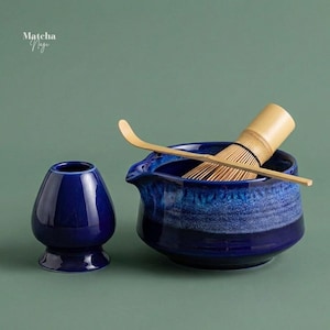 Puede incluir: Un juego de té matcha con un cuenco de cerámica azul oscuro, un batidor y una cuchara de bambú. El juego incluye un recipiente de cerámica azul más pequeño a juego. El texto "Matcha Nagi" está en la esquina superior izquierda.