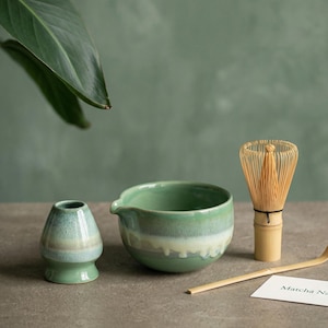 May include: A matcha tea set comprising a green ceramic bowl, a whisk, a scoop, and a small ceramic container. The set is displayed on a neutral surface with a green backdrop and a leaf.