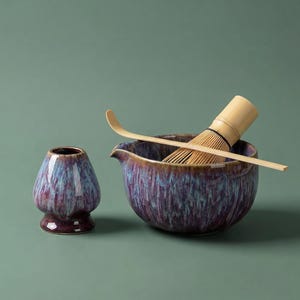 May include: A matcha tea set comprising a purple and blue glazed ceramic bowl, a bamboo whisk, a bamboo scoop, and a small ceramic container. The set is designed for preparing matcha tea. The background is a solid green.