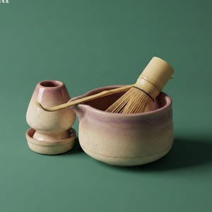May include: A matcha tea set comprising a ceramic bowl, whisk, scoop, and a small ceramic container. The bowl and container feature a beige and pink gradient. The whisk is bamboo. The text "Matcha Nagi" is in the upper left corner.