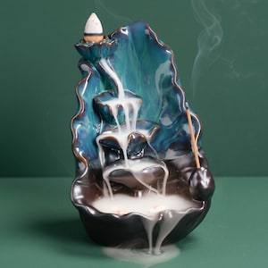 May include: A ceramic incense burner in a waterfall design. The burner is teal and brown with a glossy finish. White smoke flows from the top, creating a visual effect. An incense cone sits at the top.