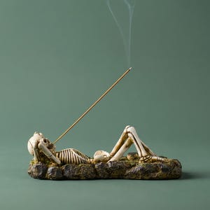 Può includere: Un portaincenso decorativo con una figura di scheletro reclinato. Lo scheletro è appoggiato su una base rocciosa strutturata, con un bastoncino d'incenso in bocca, che emette fumo. La tavolozza dei colori comprende beige, marrone e verde.