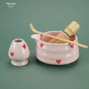 Op de afbeelding: Een matcha-theeset met een roze keramische kom en garde, een kleine roze keramische container en een bamboelepel. De kom en container zijn versierd met rode hartvormen. De tekst "Matcha Nagi" staat in de linkerbovenhoek.