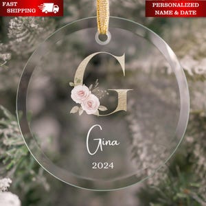 May include: Circular clear glass Christmas ornament with a gold letter "G" and the name "Gina" in white script. The ornament has a floral design with pink roses and the year "2024". A gold ribbon is attached for hanging.