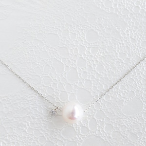 May include: A delicate necklace featuring a large, lustrous white pearl pendant. A small, sparkling diamond accent is positioned near the pearl. The necklace is crafted from a fine, silver-coloured chain, creating a minimalist and elegant design.