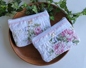 Puede incluir: Dos neceseres acolchados blancos con estampados florales en rosa, morado y verde. Cada neceser tiene un ribete de encaje, una cremallera rosa y un pequeño lazo. Los neceseres se exhiben en una bandeja de madera.