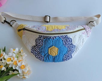 Puede incluir: Una riñonera en forma de media luna confeccionada en tela acolchada con un diseño de patchwork de patrones florales y geométricos en azul, amarillo y blanco. El bolso incluye una correa de lona beige ajustable con herrajes metálicos y cremallera.