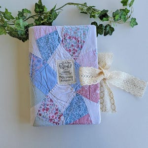 May include: A rectangular fabric-covered journal featuring a patchwork design of blue, pink, and white floral and gingham patterns. A central cream-colored fabric label displays the text God is in control and His timing is perfect. A cream lace ribbon is tied around the side.