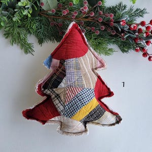 May include: A handmade Christmas tree ornament. The tree is made of patchwork fabric in various patterns and colors, including red, blue, yellow, and plaid. The edges are trimmed with a cream-colored, fluffy material. The number "1" is visible on the right side.