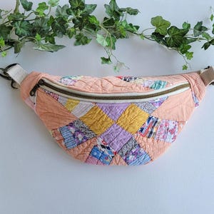May include: A crescent-shaped bum bag constructed from quilted fabric featuring a peach-coloured border and a central patchwork panel. The patchwork consists of various small squares in yellow, lavender, blue, and white patterns. It has a metal zip.