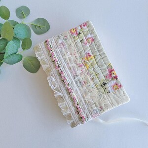 May include: A rectangular fabric book cover featuring a quilted design with vertical panels of floral print fabric in shades of pink, yellow, and green, accented with white lace trim and a decorative ribbon of tiny pink rosebuds.
