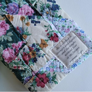 May include: A quilted fabric item with a floral pattern in shades of pink, purple, green, and cream. A small patch with text reads "She is clothed in strength and dignity and she laughs without fear of the future. Proverbs 31:25."