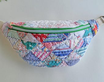Puede incluir: Un bolso de cintura acolchado con un diseño de patchwork. La bolsa presenta una variedad de patrones de tela en tonos de rojo, azul, rosa y verde. Tiene una cremallera verde y una correa color crema.