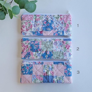 May include: Three quilted fabric zipper pouches in pink, blue, and green. Each pouch features a patchwork design with floral patterns. The pouches are rectangular and have white zips.