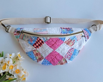 Puede incluir: Una riñonera en forma de media luna confeccionada con una colcha de retazos que presenta varios cuadrados de tela coloridos en tonos rosa, azul, rojo y blanco. El bolso incluye una correa ajustable de tela beige con herrajes metálicos.