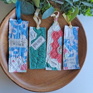 May include: Four handmade fabric bookmarks with various patterns and text. The bookmarks are in a wooden bowl. The first bookmark has blue and white floral fabric with the text "spread her wings". The second is green with the text "peace". The third is red and white with the text "vintage". The fourth is blue and white with the text "love you".