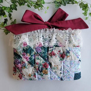 May include: A quilted fabric pouch with a burgundy bow and white lace trim. The pouch features a patchwork design with floral patterns in shades of green, pink, and cream. The bow is tied at the top, adding a decorative element.