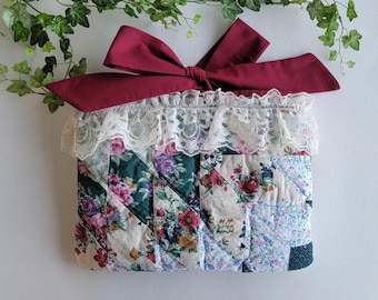 Puede incluir: Una bolsa de tela acolchada con un lazo burdeos y ribete de encaje blanco. La bolsa presenta un diseño de patchwork con estampados florales en tonos verdes, rosas y crema. El lazo está atado en la parte superior, añadiendo un elemento decorativo.