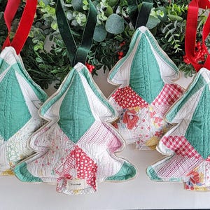 May include: A collection of Christmas tree-shaped ornaments. Each ornament is crafted from patchwork fabric in shades of green, white, and red, with a quilted texture. Red and green velvet ribbons are attached for hanging.
