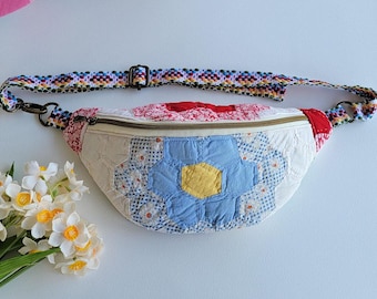 Puede incluir: Una riñonera confeccionada en tela acolchada con un diseño de patchwork de patrones florales hexagonales en azul, amarillo, rojo y blanco. El bolso incluye un cierre de cremallera metálico y una correa tejida ajustable con un patrón geométrico.