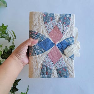 May include: A quilted journal with a patchwork design. The cover features a cream background with blue plaid, pink diamond, and floral patterned fabric accents. A cream lace ribbon is tied to the side.