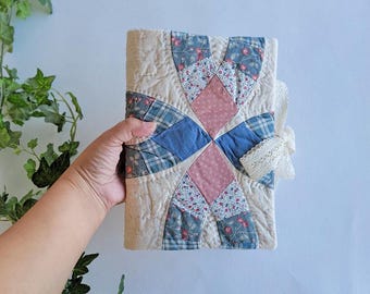 Puede incluir: Un diario acolchado con un diseño de patchwork. La cubierta presenta un fondo crema con cuadros azules, diamantes rosas y acentos de tela estampada floral. Una cinta de encaje crema está atada al costado.
