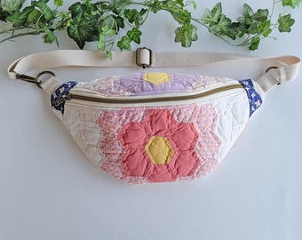Puede incluir: Una riñonera acolchada con un diseño de patchwork de patrones florales hexagonales en tonos rosa, amarillo, lavanda y blanco. El bolso incluye una correa ajustable de color beige con herrajes de metal bronce y cierre de cremallera.