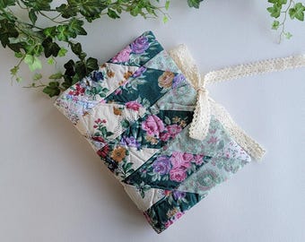 Puede incluir: Una funda de libro acolchada con un estampado floral en tonos verdes, rosas y crema. La funda está atada con una cinta de encaje color crema. El diseño presenta un estilo patchwork con varias telas florales.