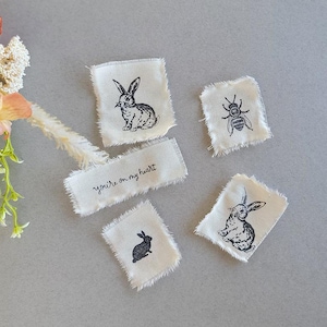 May include: Five small, white fabric squares with black ink illustrations. The squares feature a bee, two rabbits, and the phrase "you're on my heart".