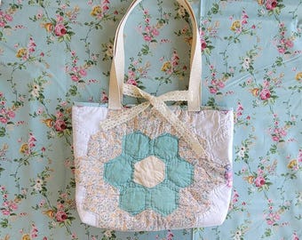 Puede incluir: Un bolso tote hecho a mano con un diseño de patchwork floral. El bolso presenta una gran flor turquesa y blanca en el centro, con un lazo color crema. Las asas son de una tela resistente de color blanquecino. El fondo es un estampado floral azul claro.