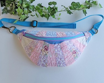Puede incluir: Una riñonera en forma de media luna con un diseño de patchwork acolchado en tonos rosa, lavanda y blanco con patrones florales. Incluye una correa ajustable de tela azul brillante, un cierre de cremallera azul a juego y anillas de metal.