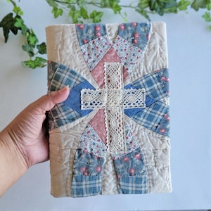 May include: A handmade, quilted book cover with a patchwork design. The cover features a central cross made of cream-coloured lace, surrounded by sections of pink floral, blue plaid, and beige fabric. The cover is held in a hand.