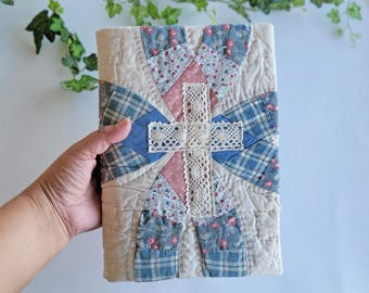 Puede incluir: Una funda de libro acolchada hecha a mano con un diseño de patchwork. La funda presenta una cruz central hecha de encaje color crema, rodeada de secciones de tela floral rosa, cuadros azules y beige. La funda se sostiene en una mano.