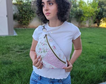 Puede incluir: Un bolso de cintura acolchado con un estampado floral y un panel rosa. El bolso tiene cierre de cremallera y una correa. El bolso es blanco con diseños florales rosas y verdes. La correa se sujeta con un cierre metálico.
