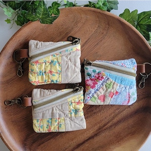 May include: Three small, square, quilted zipper pouches with attached brown leather key ring clips. Two pouches feature a yellow floral pattern with beige panels, while one pouch displays a multi-colored patchwork floral and geometric print.