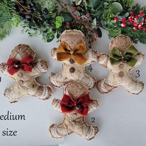 May include: Four medium-sized, handmade gingerbread man ornaments with fabric bodies and lace trim. Each has a different colored bow: red, gold, and green. The ornaments are decorated with button accents and are arranged on a white surface with greenery.