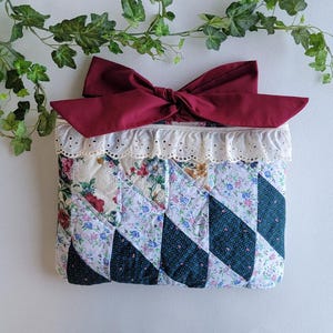 May include: A quilted clutch with a burgundy bow. The clutch features a patchwork design with floral and geometric patterns in shades of white, blue, and green. The top edge is trimmed with white lace.