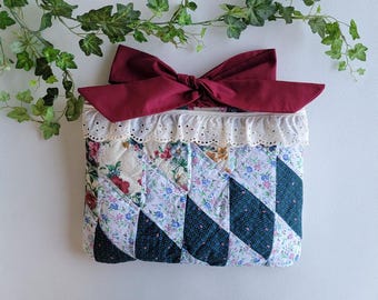 Puede incluir: Un bolso de mano acolchado con un lazo burdeos. El bolso presenta un diseño de patchwork con estampados florales y geométricos en tonos blancos, azules y verdes. El borde superior está adornado con encaje blanco.