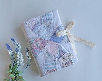 Puede incluir: Una funda de libro acolchada hecha a mano con un diseño de patchwork en colores pastel. La funda está atada con una cinta color crema. El texto "God is love" es visible. Una ramita de lavanda está en el fondo.