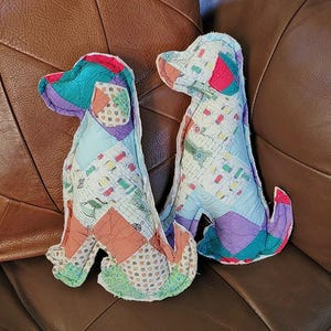 May include: Two handmade dog-shaped stuffed toys. Each dog is crafted from a patchwork of colorful fabric scraps, featuring shades of blue, green, red, and purple. The dogs are sitting upright, showcasing their unique, quilted design. They are placed on a brown leather surface.