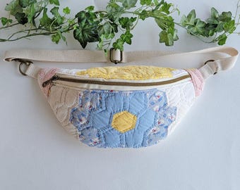 Puede incluir: Una riñonera en forma de media luna confeccionada en tela acolchada con un diseño de patchwork de patrones hexagonales florales en azul, amarillo y blanco. El bolso incluye una correa beige ajustable con herrajes metálicos y cremallera.